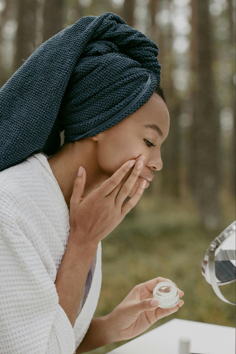 A woman in a robe with a towel on her hair applies a product to her face.