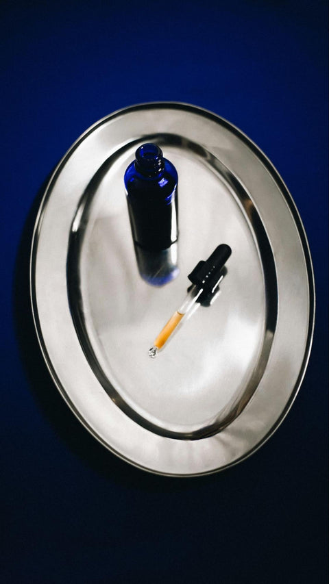 A blue dropper bottle sits on a platter with a dropper filled with serum beside it.