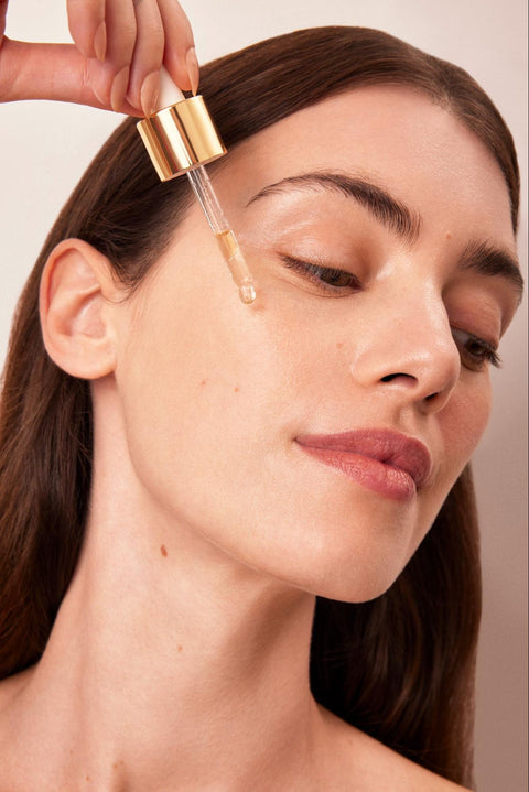 A woman uses a dropper to dispense serum onto her cheek.